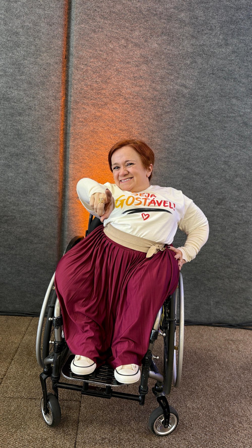 Katya Hemelrijk, sorridente, aponta para você em sua cadeira de rodas, vestindo uma camiseta com a frase 'Seja Gostável'